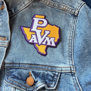 May include: A light blue denim jacket with a patch featuring the letters "P A V M" in white and purple, set against a yellow Texas-shaped background. The jacket has a buttoned pocket and a copper-colored button.