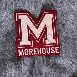 May include: Embroidered patch featuring a large white "M" within a maroon border. The word "MOREHOUSE" is stitched in white at the bottom. The patch is rectangular and has a white border. The patch is on a denim background.