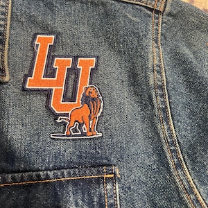 May include: A blue denim jacket with an orange and black embroidered patch featuring the letters "LU" and a lion.