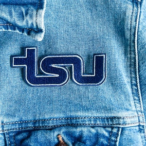 May include: A blue and white embroidered patch with the letters "TSU" on a denim jacket.