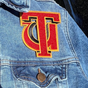 May include: A red and yellow embroidered patch with the letter "T" on a blue denim jacket.