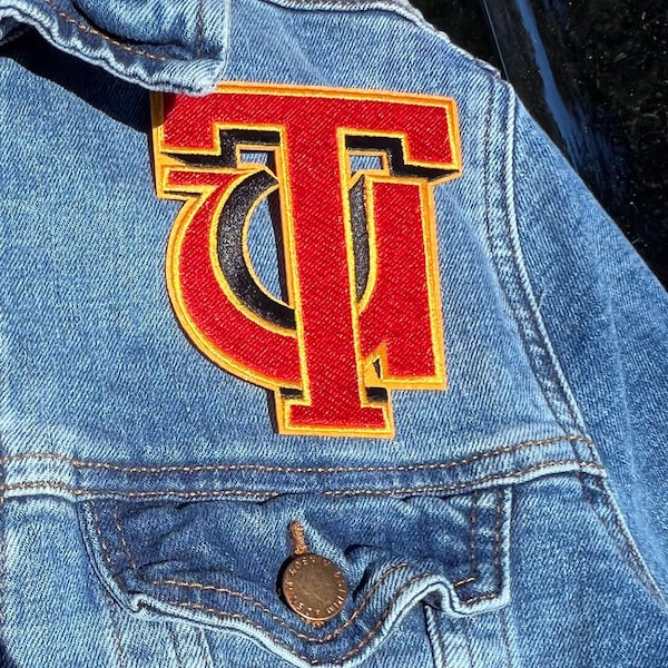 Op de afbeelding: Een rood en geel geborduurd patch met de letter "T" op een blauw denim jack.