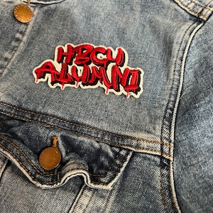 May include: Close-up of a light blue denim jacket with a patch that reads "HBCU ALUMNI" in red and black. The jacket has gold-colored buttons and visible stitching.