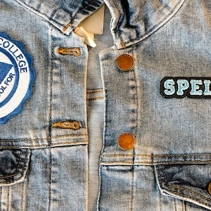 Op de afbeelding: Een blauwe denimjas met twee patches. De linker patch is een ronde patch met de tekst "Spelmen College" en "Our Whole School For Christ" rond een blauwe driehoek met een ster. De rechter patch is een rechthoekige patch met de tekst "Spelmen" in blauwe letters.