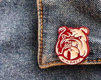 Op de afbeelding: Een rode emaille pin met een wit bulldoghoofd en de tekst "TAMU" in witte letters. De pin is op een blauw denim jack.