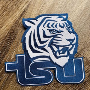 Peut inclure: Écusson brodé représentant une tête de tigre blanche et bleue, avec les lettres "TSU" en bleu avec un bord blanc. L'écusson est posé sur une surface en bois.