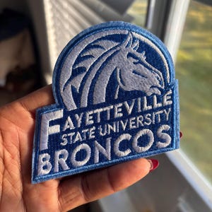 May include: A blue and white embroidered patch featuring a horse head and the words "FAYETTEVILLE STATE UNIVERSITY BRONCOS". The patch has a rectangular base with a curved top, and is likely a collegiate emblem.