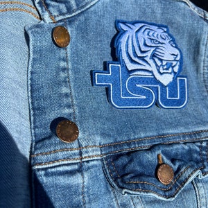 May include: A blue denim jacket with a blue and white embroidered tiger and the letters "TSU" on the left side of the jacket. There are two gold buttons on the jacket.