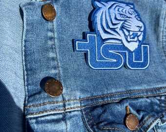 Peut inclure: Une veste en jean bleue avec un tigre brodé bleu et blanc et les lettres "TSU" sur le côté gauche de la veste. Il y a deux boutons dorés sur la veste.