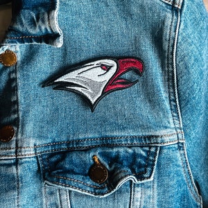 May include: A blue denim jacket with a white and red embroidered eagle patch on the front. The eagle has a red beak and a white eye.