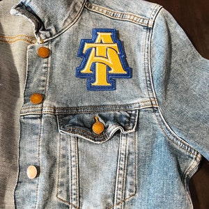 May include: Light blue denim jacket with a patch featuring the letters "AT" in yellow and blue. The jacket has a classic design with button closures and a chest pocket. The buttons are a copper color.