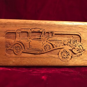 May include: A rectangular wooden box with a detailed carved image of a vintage car. The car is a classic model with visible wheels, headlights, and a detailed body. The wood has a natural grain and a warm brown tone.