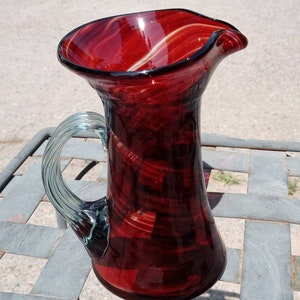 May include: A hand-blown glass pitcher with a deep red hue and a clear, twisted handle. The pitcher has a flared rim and a slightly curved spout. The glass has a marbled appearance with streaks of lighter red and white. The pitcher is a decorative item.