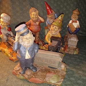 May include: A collection of whimsical gnome figurines, each with unique attire and expressions. The gnomes are posed around a miniature train, with details including a conductor, engineer, and passengers. The color palette includes reds, greens, and browns.
