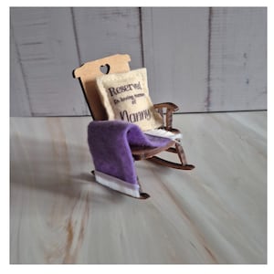 May include: A miniature wooden rocking chair with a heart-shaped cutout in the back. A cream-colored pillow with the text "Reserved in loving memory of Nanny" is placed on the chair. A purple blanket is draped over the chair.