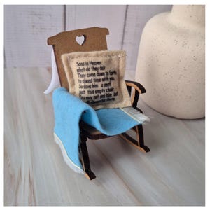 May include: A miniature wooden rocking chair with a light blue blanket draped over the seat. A small beige pillow with the text "Sons in Heaven what do they do? They come down to Earth to spend time with you So save him a seat just this empty chair you may not see him but he always be there" is on the seat.