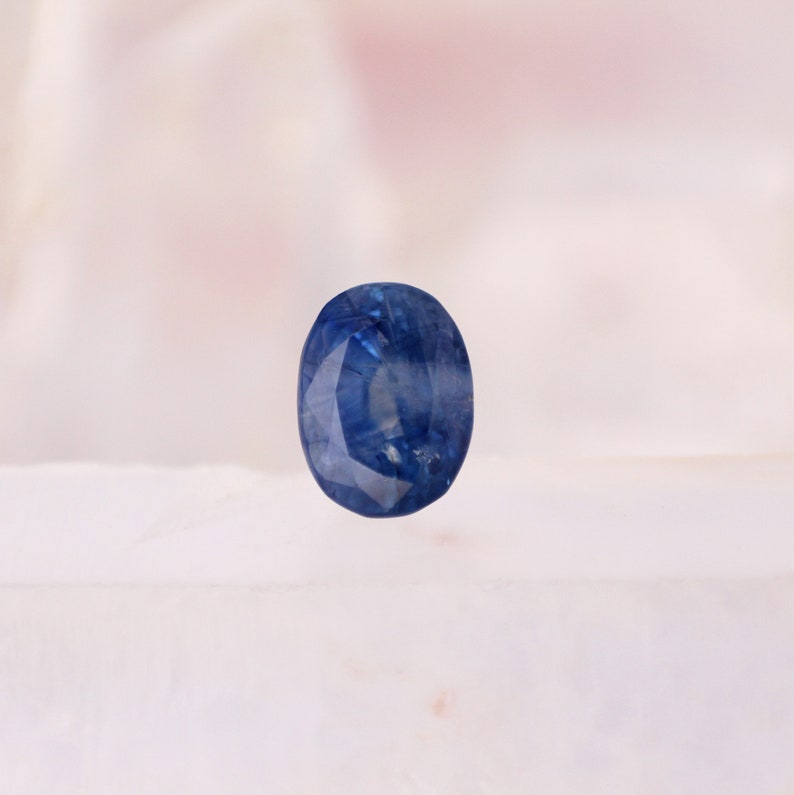 May include: A faceted, oval-shaped sapphire gemstone. The sapphire is a deep, vibrant blue color, with visible internal structures. The gemstone is set against a soft, blurred background, highlighting its color and clarity. The gem is a classic choice for jewelry.