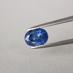 May include: A faceted, oval-shaped blue gemstone is held by a pair of silver tweezers against a light gray background. The gemstone has a brilliant, reflective surface, showcasing its facets and internal structure. The gem's color is a deep, saturated blue.