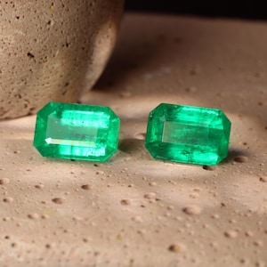 May include: Two vibrant, emerald-cut emerald gemstones. The gems are a rich, translucent green, with visible internal structures. The stones are displayed on a textured, light brown surface, showcasing their brilliance.