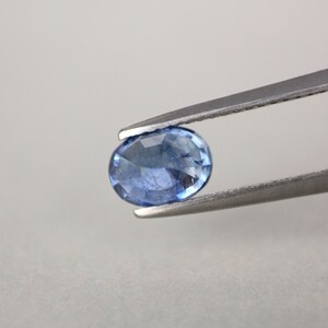 May include: A faceted, oval-shaped blue gemstone held by a pair of silver tweezers. The gem has a light blue hue with visible internal facets, reflecting light. The background is a plain, light gray.