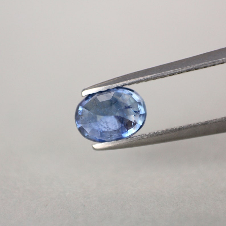 May include: A faceted, oval-shaped blue gemstone held by a pair of silver tweezers. The gem has a light blue hue with visible internal facets, reflecting light. The background is a plain, light gray.