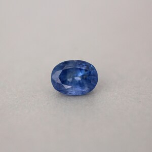May include: An oval-shaped, faceted sapphire gemstone. The gem is a deep, saturated blue color with internal light reflections. The stone is set against a neutral, light gray background, highlighting its clarity and brilliance.