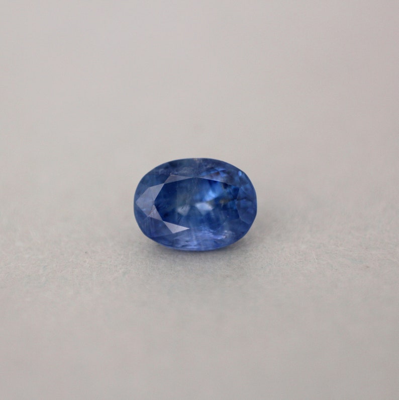 May include: An oval-shaped, faceted sapphire gemstone. The gem is a deep, saturated blue color with internal light reflections. The stone is set against a neutral, light gray background, highlighting its clarity and brilliance.