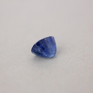 May include: A faceted, oval-shaped sapphire gemstone. The sapphire is a deep, rich blue color with visible internal structures. The gemstone is set against a neutral background, highlighting its color and cut.