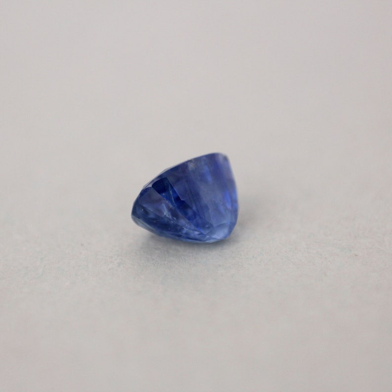 May include: A faceted, oval-shaped sapphire gemstone. The sapphire is a deep, rich blue color with visible internal structures. The gemstone is set against a neutral background, highlighting its color and cut.