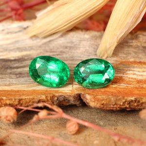May include: Two oval-shaped emerald gemstones, a vibrant green color, set against a rustic wooden background.