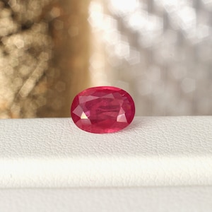 May include: A vibrant red oval-shaped gemstone, likely a ruby, is displayed on a white surface.