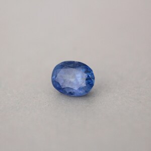 May include: A faceted, oval-shaped sapphire gemstone. The gem is a deep, vibrant blue color with a polished surface. The sapphire is set against a neutral background, highlighting its color and clarity.