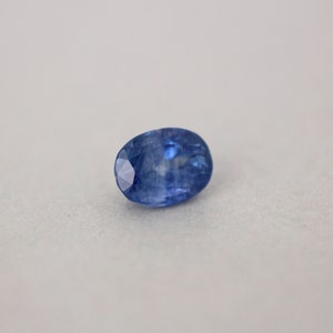 May include: A faceted, oval-shaped blue gemstone. The gem has a deep, saturated blue color with a polished surface. The facets catch the light, creating a sparkling effect. The gemstone is set against a neutral background.