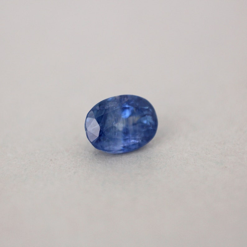 May include: A faceted, oval-shaped blue gemstone. The gem has a deep, saturated blue color with a polished surface. The facets catch the light, creating a sparkling effect. The gemstone is set against a neutral background.