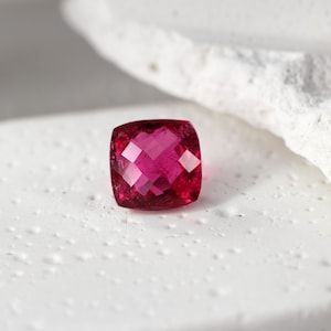 Peut inclure: Une pierre précieuse rose vif de taille coussin, présentant un design multi-facettes. La gemme est exposée sur une surface blanche.