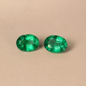 May include: Two oval-shaped emerald green gemstones on a beige background.