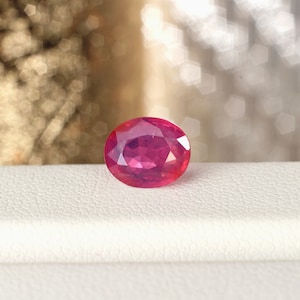 May include: A vibrant, oval-cut ruby gemstone. The gem is a deep, saturated red color, with facets that catch the light. The ruby is set against a white surface, with a blurred, bokeh background.