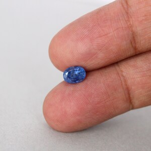 May include: A small, oval-shaped, blue gemstone. The gem is held between two fingers against a white background. The stone has a polished surface and a vibrant, saturated color.