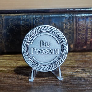 May include: A silver-toned round token with a rope-like border and the words "Be Present" in a circle in the center. The token is on a clear plastic stand.
