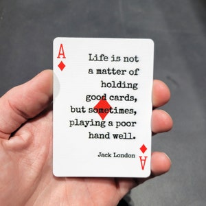 May include: A white playing card with a red ace of diamonds on the front and back. The card features a quote by Jack London: "Life is not a matter of holding good cards, but sometimes, playing a poor hand well."