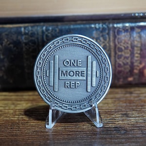 May include: A silver-toned coin with a dumbbell design and the words "ONE MORE REP" engraved on it. The coin is on a clear plastic stand.