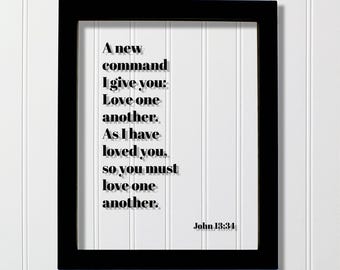 May include: A black rectangular frame containing a white background with black text that reads: A new command I give you: Love one another. As I have loved you, so you must love one another. John 13:34. The text features a subtle drop shadow effect.
