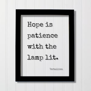May include: A black rectangular frame containing a white print with the black typewriter-style text: Hope is patience with the lamp lit. The name Tertullian is printed in smaller black text at the bottom right corner of the white background.