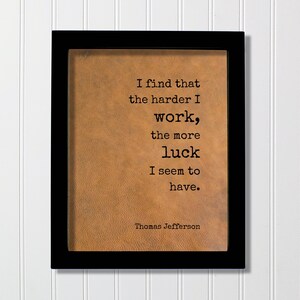 May include: A rectangular black frame containing a tan, textured background with the black printed text: I find that the harder I work, the more luck I seem to have. Thomas Jefferson. The quote is centered in a typewriter-style font.