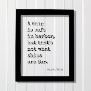 May include: A black rectangular frame containing a white print with the black typewriter-style text: A ship is safe in harbor, but that's not what ships are for. The author's name, John A. Shedd, is printed in the bottom right corner of the white area.