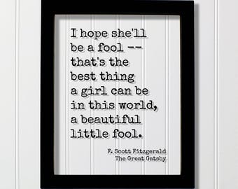 May include: A black rectangular frame containing a white print with the black typewriter-style text: I hope she'll be a fool -- that's the best thing a girl can be in this world, a beautiful little fool. F. Scott Fitzgerald, The Great Gatsby.