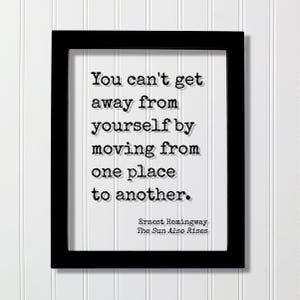 Op de afbeelding: Een zwarte rechthoekige lijst met een witte print met de zwarte tekst in typemachinestijl: You can't get away from yourself by moving from one place to another. Ernest Hemingway, The Sun Also Rises staat in de rechterbenedenhoek gedrukt.