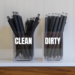 Peut inclure: Deux récipients en acrylique transparents, étiquetés "CLEAN" et "DIRTY", contiennent des stylos noirs. Les stylos sont soigneusement rangés dans chaque récipient, suggérant une organisation. Les récipients sont posés sur une étagère blanche.