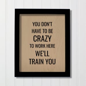 May include: A rectangular wall sign featuring a black frame and a light brown wood-grain background. The text in black capital letters reads: YOU DON'T HAVE TO BE CRAZY TO WORK HERE WE'LL TRAIN YOU. The sign has a clean, modern, and professional appearance.