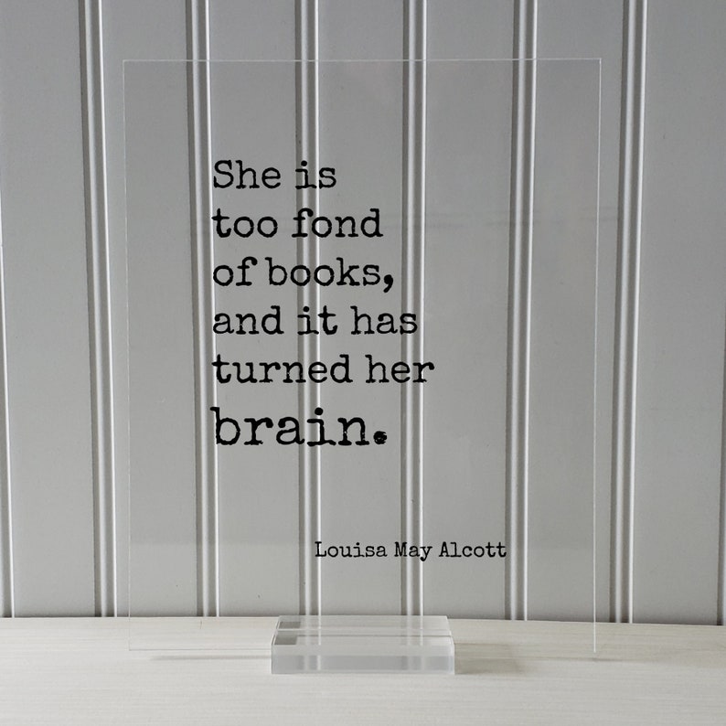 May include: A clear acrylic sign with a black quote that reads "She is too fond of books, and it has turned her brain." The quote is attributed to Louisa May Alcott.